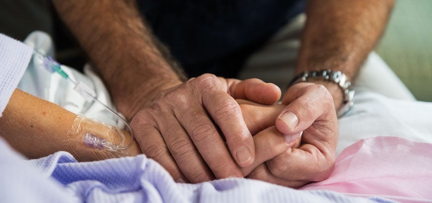 A closeup of two people holding hands with one of them being a patient lying in a hospital bed