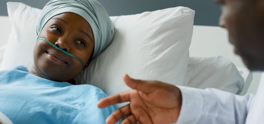 A woman lays in a hospital bed wearing a head wrap and nasal cannula while speaking to a doctor<br>  