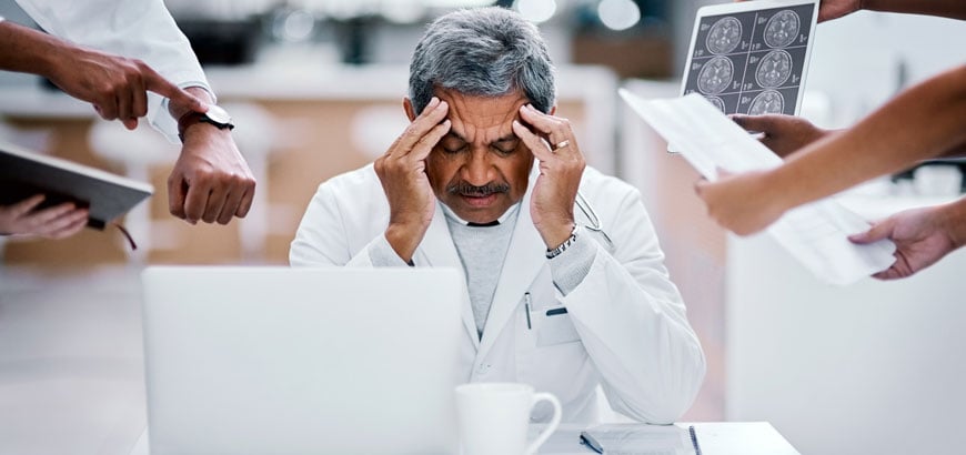 A doctor sits at his desk overwhelmed with his hands to his head as people try to talk to him and show him things